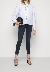 Camisa blanca de manga larga, cerrada con botones, sobre una blusa de rayas en blanco y negro. Jeans oscuros ajustados con borde deshilachado, acompañados de tacones negros con tiras.