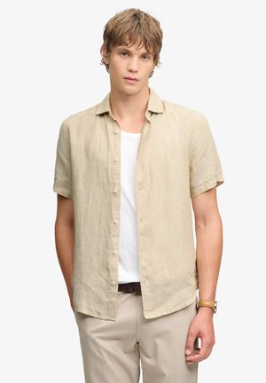 Junger Mann trägt ein beiges kurzärmeliges Hemd über einem weißen T-Shirt mit beigen Hosen und einer braunen Lederuhr, steht vor einem schlichten Hintergrund.