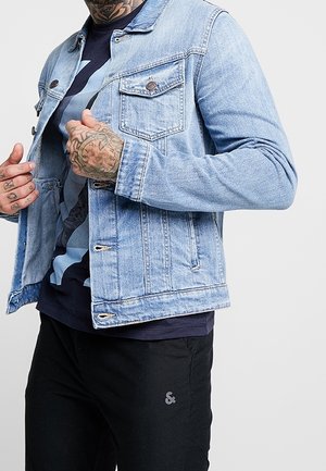 Person, der eine hellblaue Jeansjacke über einem schwarzen Grafik-T-Shirt und schwarzen Hosen trägt, zeigt tätowierte Hände, die die Jacke anpassen.