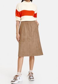 Nainen, jolla on yllään valkoinen ja oranssi raidallinen neule, ruskea polvimittainen hame, jossa on taskut, beige sukat ja harmaa-valkoiset lenkkarit.