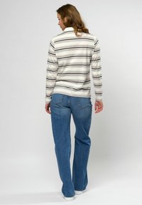 Vrouw van achteren gezien, gekleed in een wit shirt met lange mouwen en zwarte strepen, blauwe jeans met rechte pijpen en witte schoenen, tegen een effen achtergrond.