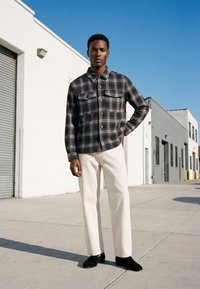 Un homme se tient dehors, portant un pantalon blanc, une chemise noire à carreaux boutonnée et des chaussures noires, devant un bâtiment blanc sous un ciel bleu clair.