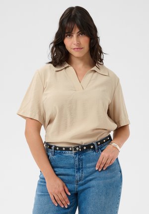 Femme aux cheveux foncés mi-longs portant un haut beige à manches courtes rentré dans un jean bleu taille haute avec une ceinture noire cloutée.