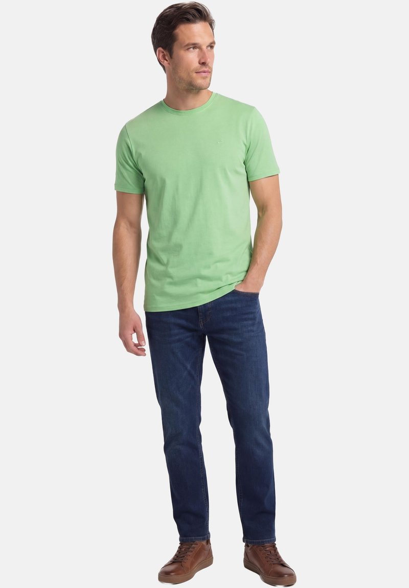 Homme debout, main gauche dans la poche, portant un t-shirt à manches courtes vert clair, un jean bleu foncé et des baskets en cuir marron, regardant vers la droite.