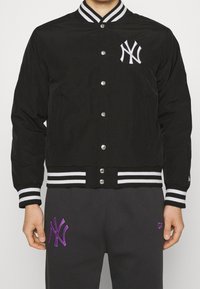 Black bomber jacket with white and black striped collar and cuffs, featuring a white logo and snap buttons on the front. Textured fabric.