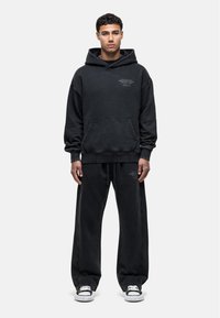 Zwarte oversized hoodie met een voorzak en logo-opdruk, gecombineerd met bijpassende relaxed-fit joggingbroek. Beide kledingstukken zijn gemaakt van een zachte, getextureerde stof.