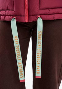 Bordó dzseki piros cipzárral és türkiz húzózsinórokkal, rajta a "WRAP YOURSELF IN STORIES" felirat, sötétbarna nadrág felett viselve.