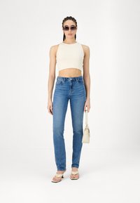 Top corto blanco, jeans ajustados azules, bolso de mano beige claro y sandalias con tiras. Las gafas de sol completan el atuendo casual.