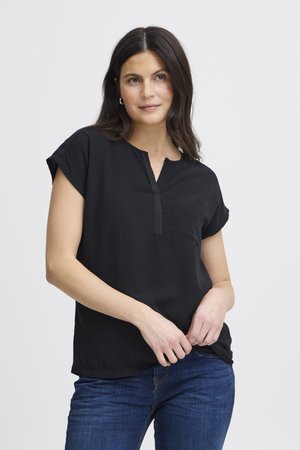 Femme aux cheveux foncés portant un chemisier noir à manches courtes et un jean bleu, debout devant un fond clair uni.