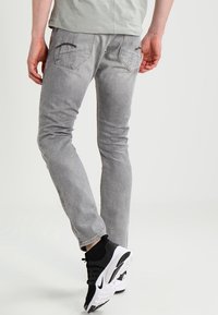 Jeans de mezclilla gris de corte ajustado, con dos bolsillos traseros y detalles de costura marrón, combinados con zapatillas deportivas negras y blancas.