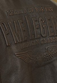 Brun läderjacka med präglad text "PME Legend" och "Legend of the Sky", med vingemblem och strukturerad korn.
