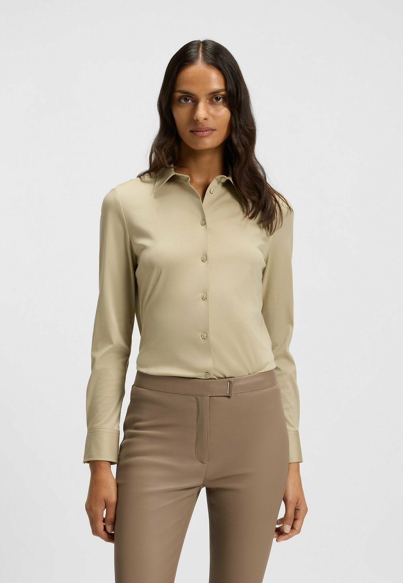 Beige Langarmhemd mit Knopfverschluss, klassischem Kragen und glattem Stoff; kombiniert mit eng sitzenden braunen Hosen mit einem eleganten Design.