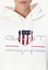 Biela mikina z bavlny s veľkým emblémom v červených, bielych a modrých detailoch, a textom "GANT AMERICAN SPORTSWEAR" nižšie.