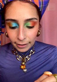 Vrouw met meerkleurige oogschaduw in blauw, groen en rood, draagt grote gouden oorbellen, gelaagde chunky kettingen en een paarse top met een bijpassende hoofddoek.