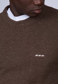 Pull en maille marron avec un col côtelé et un motif animal brodé blanc. Le tissu a un aspect texturé et une profondeur subtile.