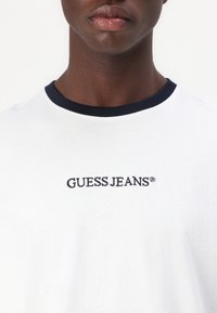 Camiseta de algodón blanca con cuello redondo azul marino, que presenta "GUESS JEANS" bordado en azul oscuro en el pecho. Textura suave.