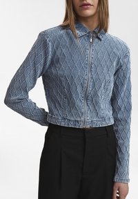 Kraftig denimjacka med diamantkviltmönster i ljusblått. Har en ståkrage och dragkedja fram. Bärs med svarta byxor.