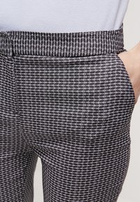 Närbild av byxor med ett svart och vitt mönster, där en hand är i framfickan, bärs tillsammans med en enkel vit topp.