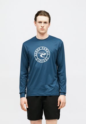 Νέος άνδρας που φορά μπλε πουκάμισο με μακρύ μανίκι και λογότυπο "Aloha Hawaii Ripcurl" και μαύρα σορτς, στέκεται μπροστά σε απλό φωτεινό φόντο.