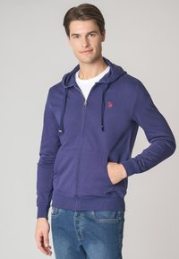 Felpa con zip blu navy in misto cotone, dotata di tasca a marsupio, cappuccio con cordino e un piccolo logo rosso sul petto. Vestibilità casual.