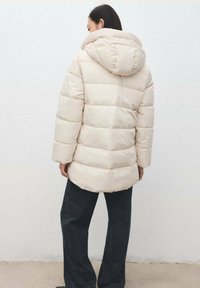 Kobieta zwrócona tyłem, ubrana w jasnobeżową pikowaną kurtkę puchową z kapturem oraz ciemne spodnie o szerokich nogawkach, na tle jednolitej ściany.