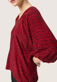 Rød løs bluse med en struktureret mønster af sorte firkanter. V-hals design med samlede ærmer og en buet nederste kant.