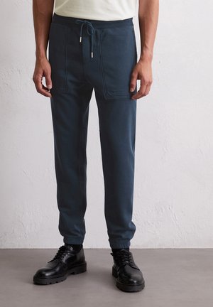 Donkerblauwe joggingbroek met een verstelbare tailleband, voorzien van voorzakken en een subtiele logo-opdruk. Glad textuur en taps toelopende boorden. Zwarte schoenen.