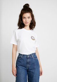Hvid bomulds t-shirt med korte ærmer, rund hals, med et cirkulært broderet logo på venstre bryst. Kombineret med højtaljede blå jeans.