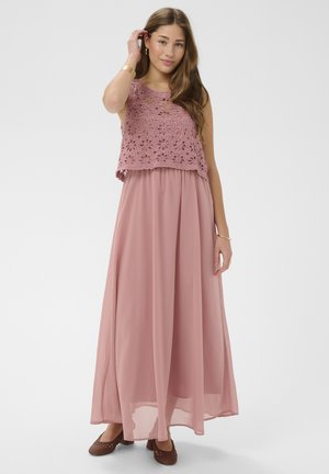 Kobieta w rękawach brzoskwiniowego różowego koronkowego topu na ramiączkach i pasującej, długiej, zwiewnej spódnicy, w brązowych plecionych balerinach, pozująca z ręką przy włosach.