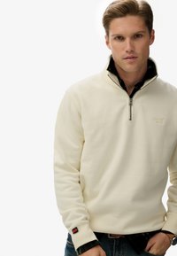 Crème-kleurige quarter-zip sweatshirt met ribgebreide manchetten en kraag, met een klein logo op de borst. Gelaagd over een geruit shirt.