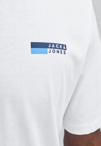 Bílý bavlněný tričko s kulatým výstřihem. Na levé části hrudi je modro-námořnické logo s textem "JACK&JONES". Hladká textura.