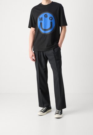 Homme portant un T-shirt noir avec un grand visage souriant bleu et les lettres "HUGO", un pantalon noir et des baskets noires avec des semelles blanches.
