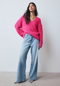 Roze grove gebreide trui met een V-hals, gecombineerd met lichtblauwe wijde jeans en zilveren puntige schoenen. Gladde vloer en neutrale achtergrond.