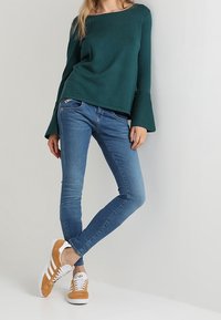 Zelený pletený svetr s volánovými rukávy, doplněný modrými džíny a béžovými semišovými teniskami s bílými pruhy. Hladká textura a přiléhavý střih.