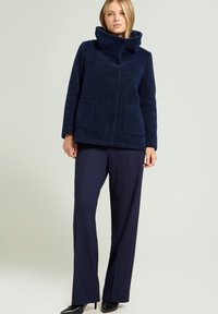 Kobieta ubrana w granatową kurtkę o fakturze z wysokim kołnierzem, w połączeniu z luźnymi ciemnymi spodniami i czarnymi szpilkami, stojąca na tle jednolitym.