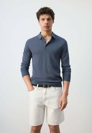 Joven de pie con las manos en los bolsillos, vestido con una camisa azul de manga larga con cuello y pantalones cortos blancos con un cinturón negro.