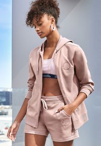 Sudadera rosa claro con cierre de cremallera y bolsillos, combinada con pantalones cortos a juego que cuentan con una cintura elástica y detalle del logo. Tejido suave y casual.