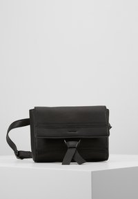 Černá kožená crossbody taška s klopou, prošíváním a předním popruhem, vystavená na bílé podstavě na šedém pozadí.