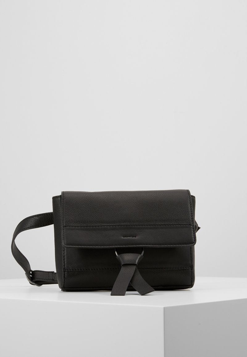 Černá kožená crossbody taška s klopou, prošíváním a předním popruhem, vystavená na bílé podstavě na šedém pozadí.