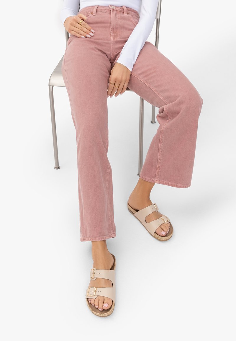 Personne assise sur une chaise en métal portant un pantalon large rose poussiére, un haut à manches longues blanc, des sandales beiges à double boucle et des bagues en or.