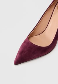 Μεχτές γόβες με μυτερή μύτη σε μπορντό σουέτ και λείο εσωτερικό επένδυση από μπεζ δέρμα σε ανοιχτό γκρι φόντο.