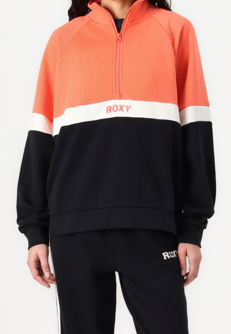 Femme portant un pull color block avec des sections corail, blanc et noir, une fermeture éclair au quart et le logo "ROXY" sur la poitrine et le pantalon.