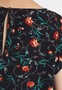 Blůza z látky s květinovým vzorem na tmavě modrém pozadí. Obsahuje červeno-oranžové květy a zelené listy, s výstřihem ve tvaru klíčové dírky a zapínáním na knoflík.