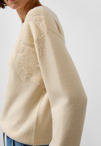 Beiger Strickpullover mit strukturiertem Sternmuster auf der oberen Brust und den Schultern, getragen von einer Person in blauer Jeans vor weißem Hintergrund.