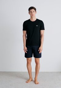Schwarzes kurzärmeliges T-Shirt mit weißem Logo auf der Brust, kombiniert mit dunkelblauen Shorts, steht auf einem hellen Teppichboden.