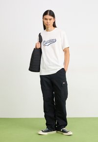 Hvit t-skjorte med blå "Converse" logo, svarte cargobukser med lommer, svart quiltet veske og svart-hvite sneakers på grønn gulv.