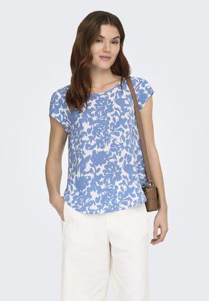 Woman wearing a blue and white floral short-sleeve top, white pants, and a brown crossbody bag, standing with one hand in pocket.