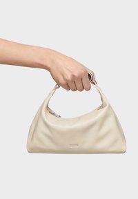 Mano sosteniendo un pequeño bolso de cuero crema suave con un asa corta y herrajes plateados sobre un fondo blanco.