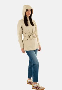 Giacca impermeabile beige con cappuccio, vita cintrata, bottoni a pressione e due tasche frontali. Abbinata a jeans blu e sneakers beige.