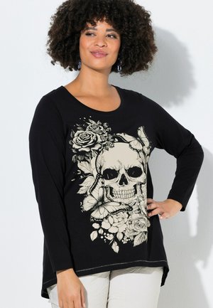 Donna con capelli ricci che indossa una maglietta nera a maniche lunghe con una grafica beige di teschio, rose e farfalle.